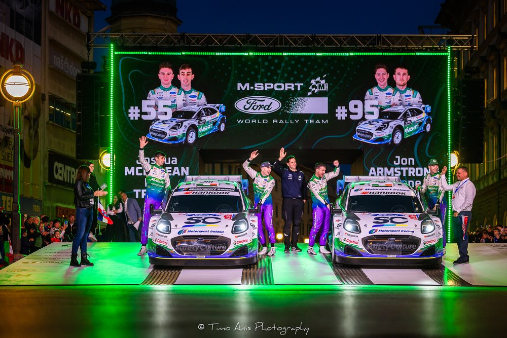 M-Sport Ford World Rally Team drivers celebrate on podium with cars #55 and #95 at night rally event.