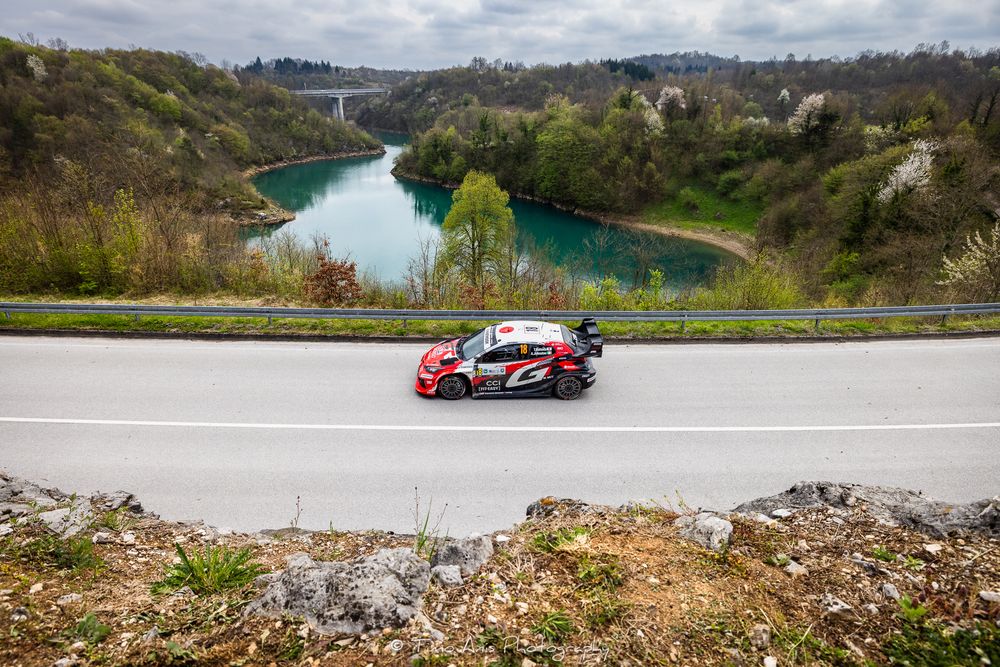 A rally car speeds along a winding road above a scenic river valley with a bridge and lush green hills in the background.