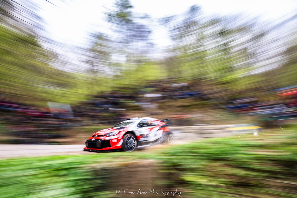 A rally car speeds through a forest stage with dynamic motion blur effect in a WRC racing competition.