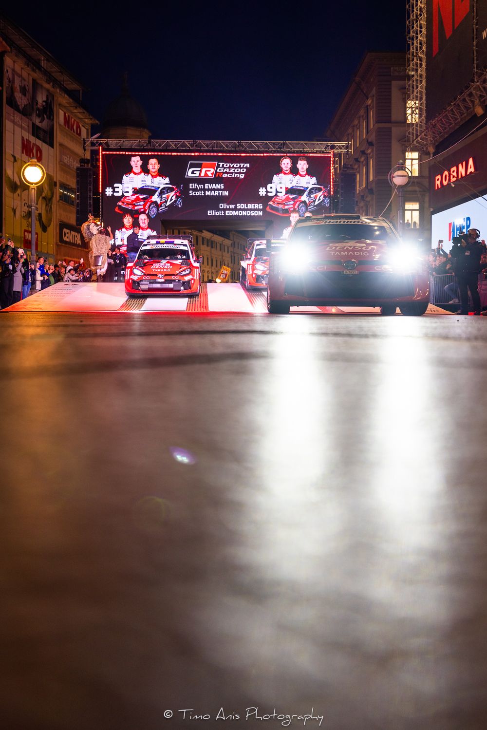 Toyota Gazoo Racing WRC rally cars on illuminated night stage with bright headlights and team display screen in city setting.