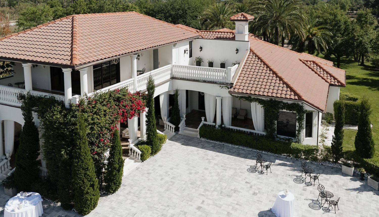 Aerial view of a luxurious white Mediterranean-style villa with red tile roof, manicured hedges, and elegant patio seating.