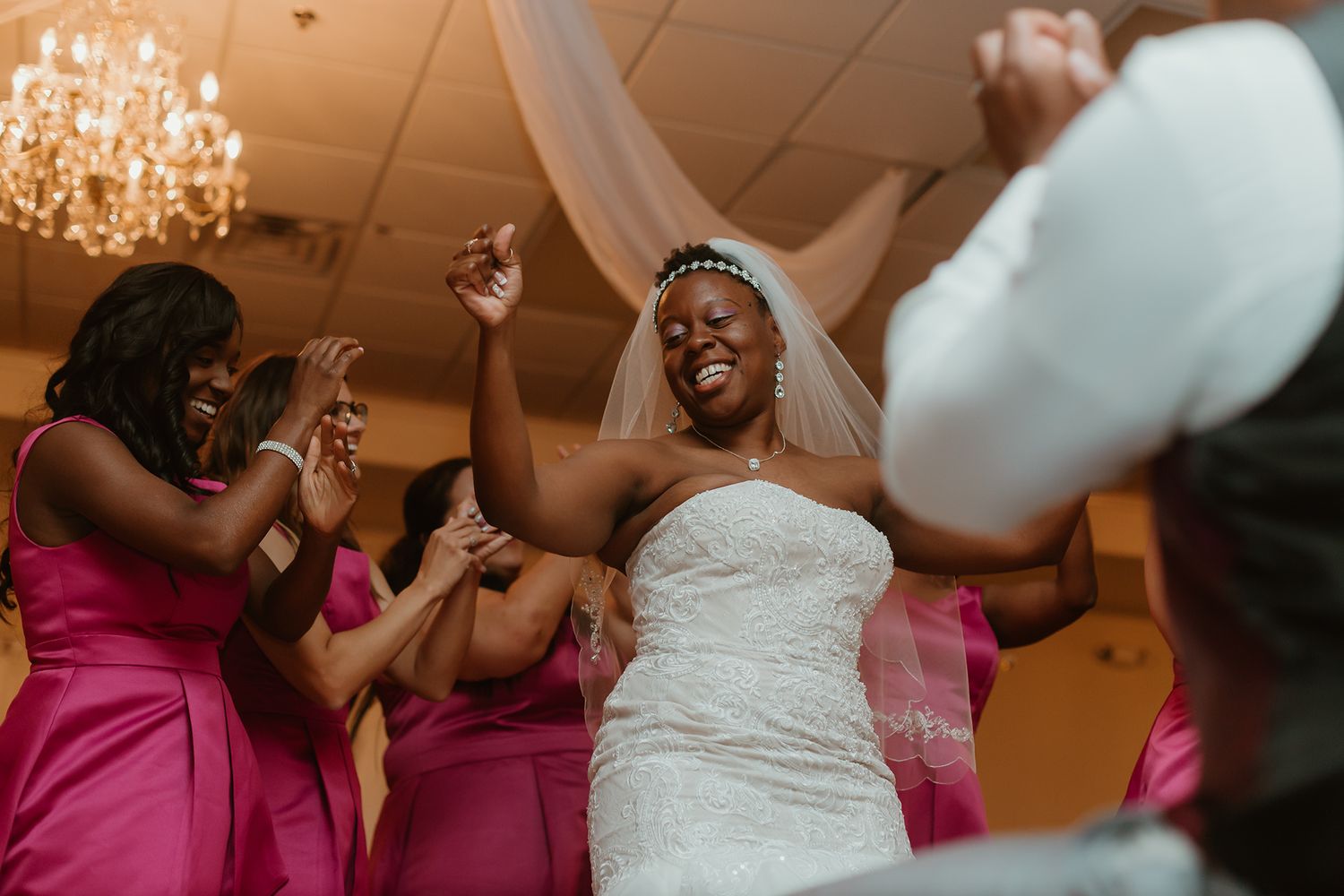 Joyful bride in white gown and veil dancing with pink-dressed bridesmaids at wedding reception.