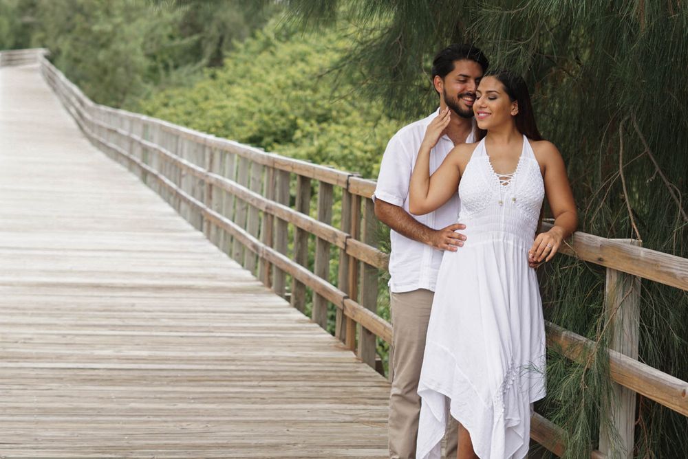Couple in white outfits embracing on a wooden bridge surrounded by lush green trees.
