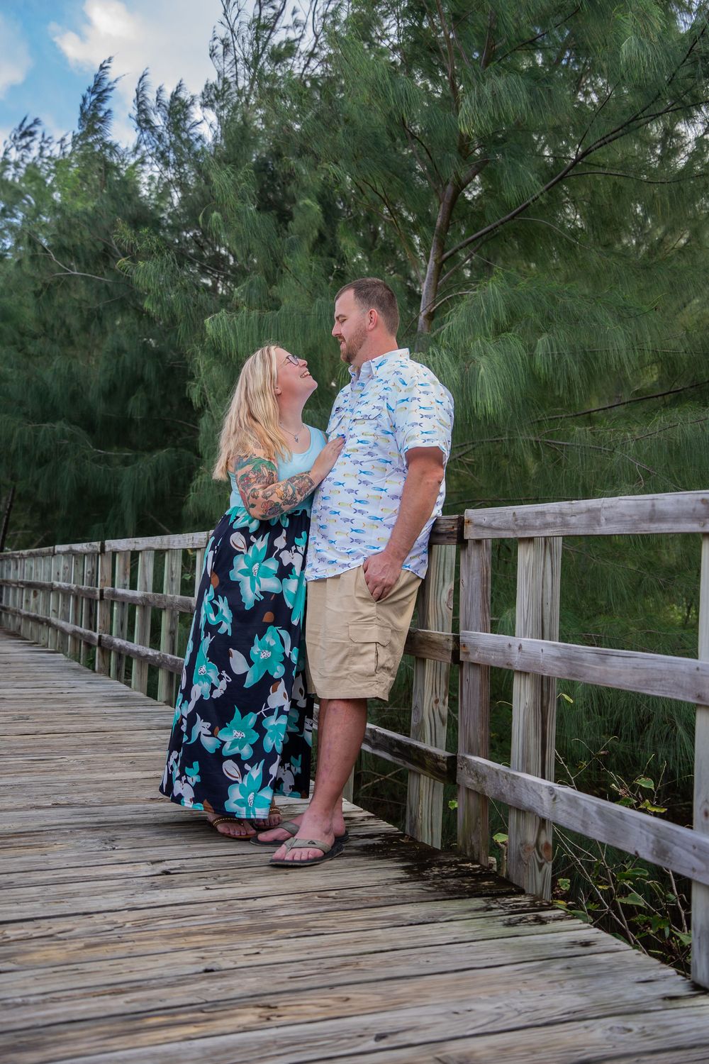 Couple holding hands on a wooden bridge surrounded by lush green trees, woman in floral dress gazing up at man.