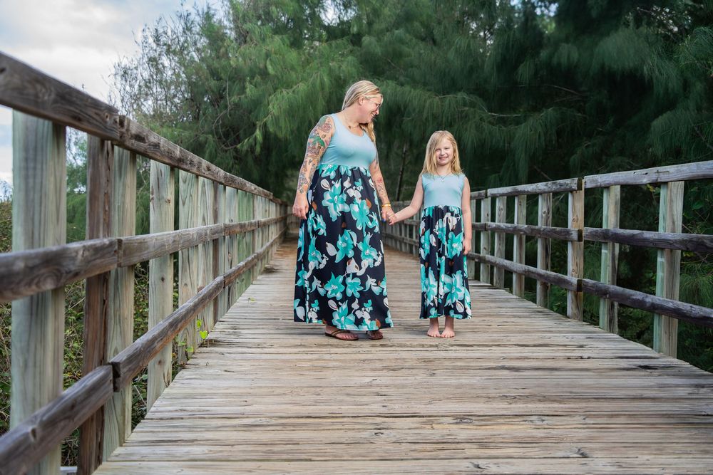 Mother and daughter in matching floral maxi dresses holding hands on a wooden bridge outdoors.