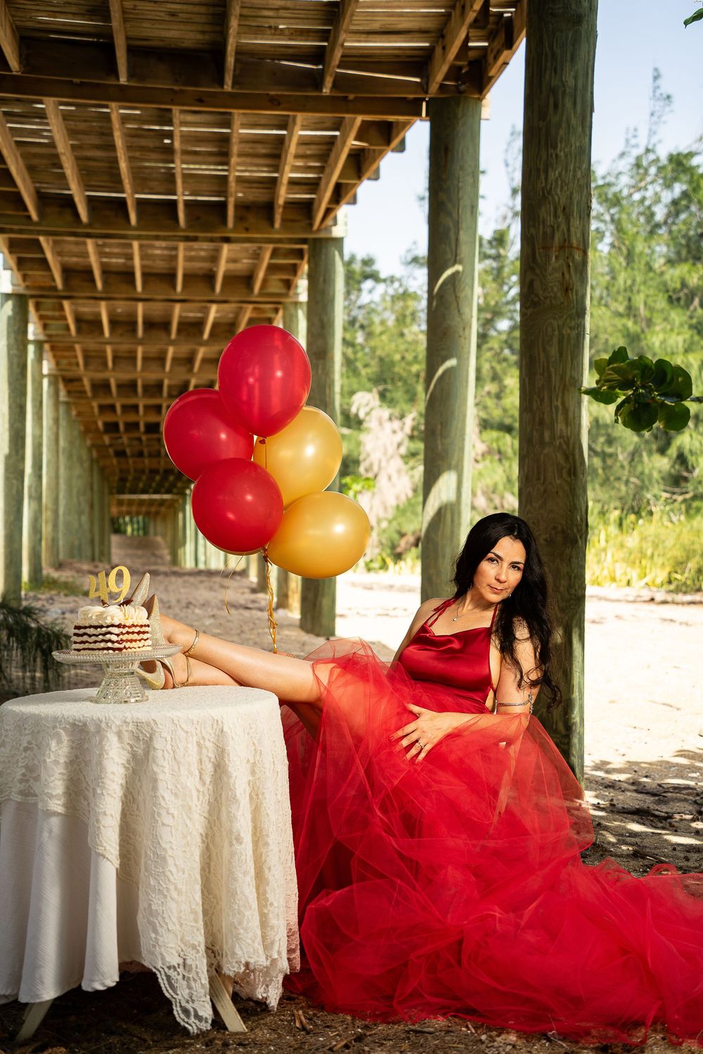 Woman in red tulle gown poses with colorful balloons beside a birthday cake at an outdoor celebration.