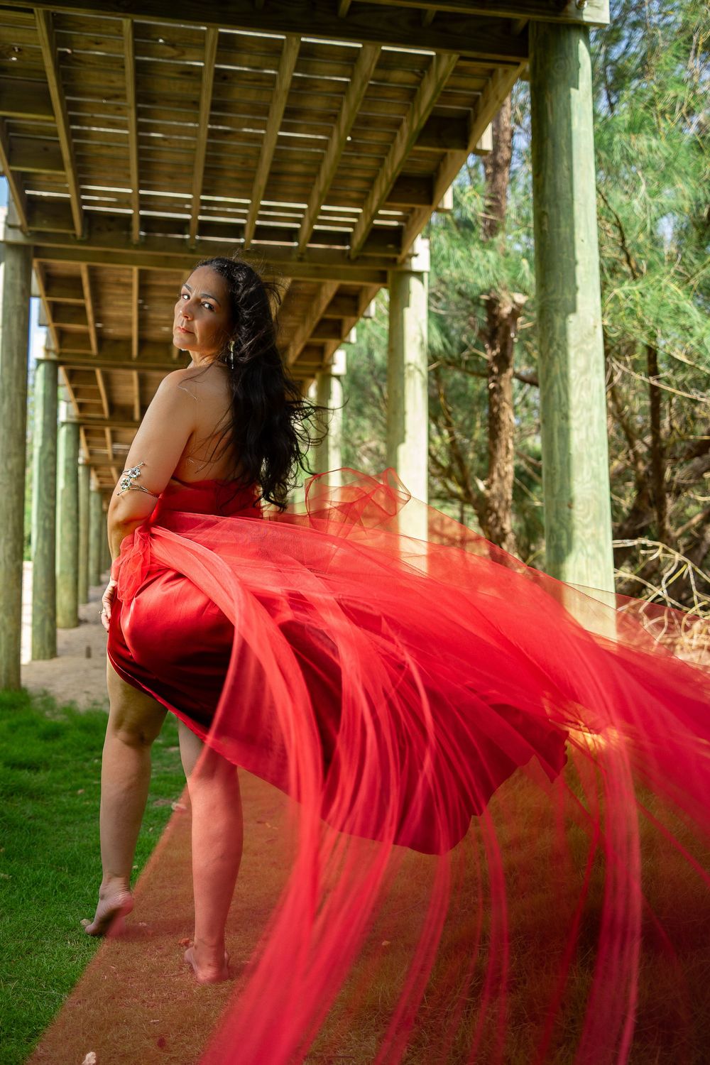 Woman in flowing red dress poses gracefully under wooden pergola surrounded by lush green trees.
