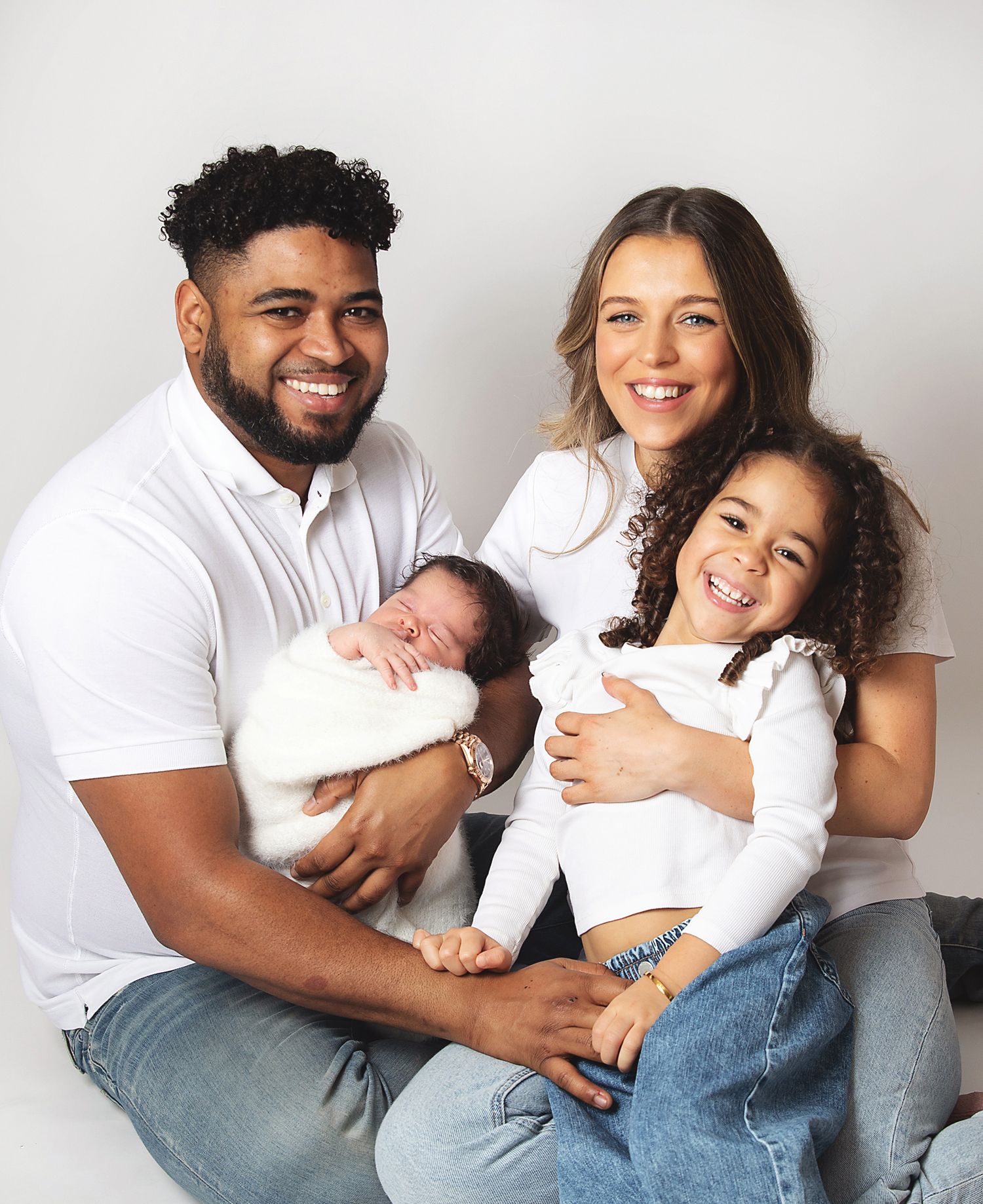 family portrait with parents holding newborn baby and young daughter, all wearing white tops and jeans. During a newborn session in London