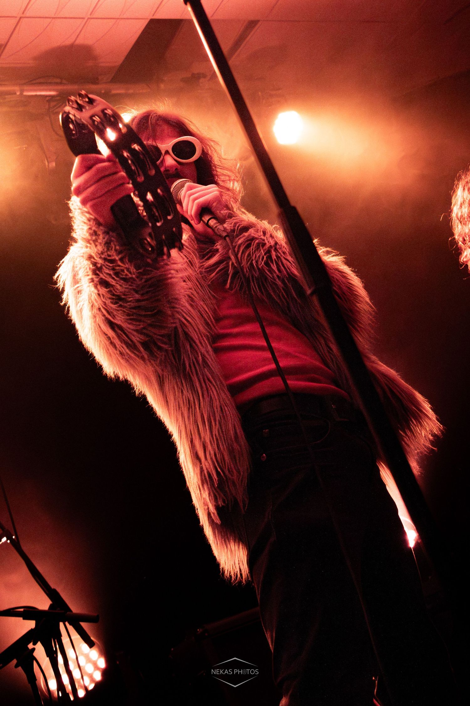 A dramatic rock performer in a fur coat and goggles holds a guitar aloft under intense red stage lighting.