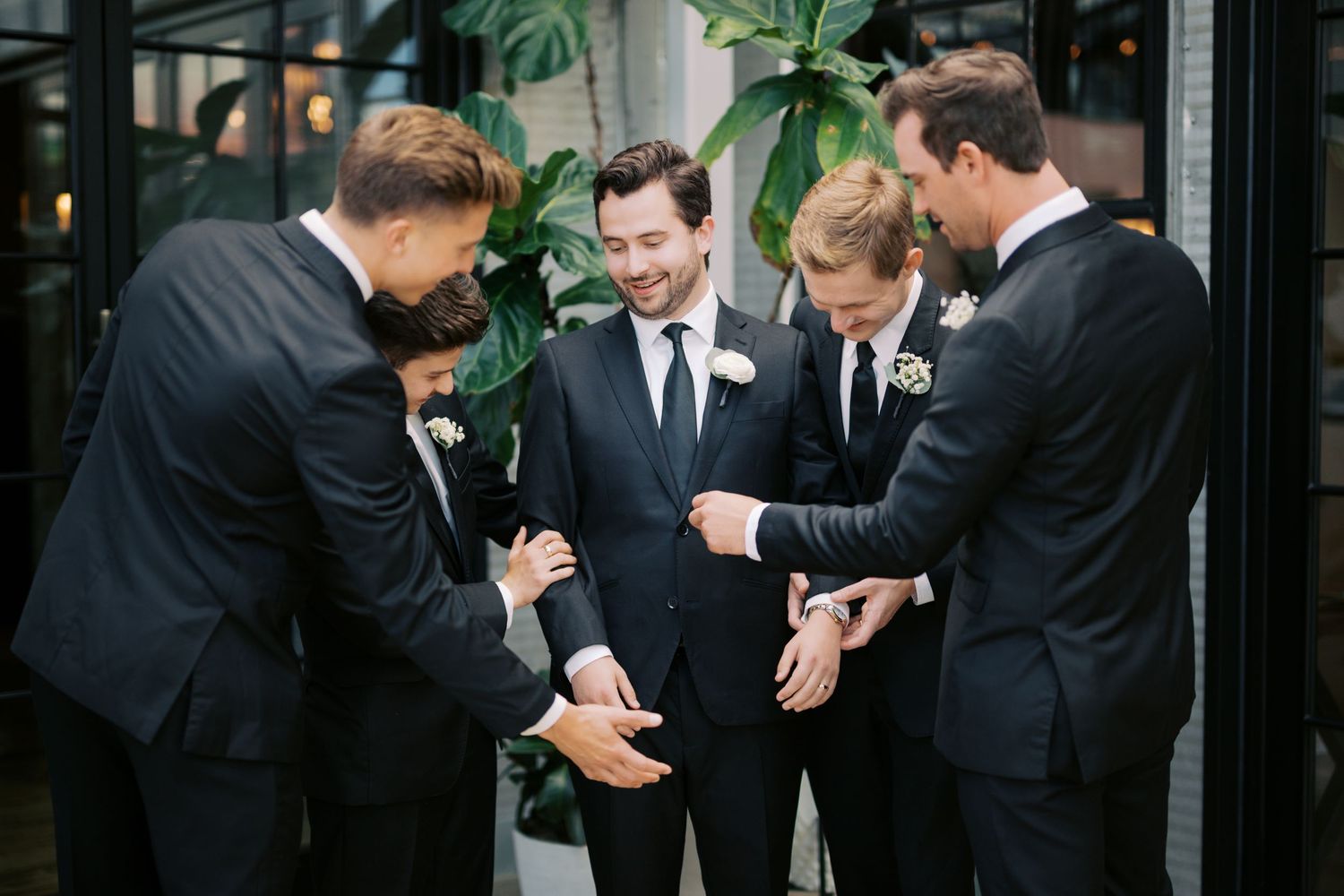 Groom and groomsmen in black suits sharing a joyful moment together before a wedding ceremony outdoors.