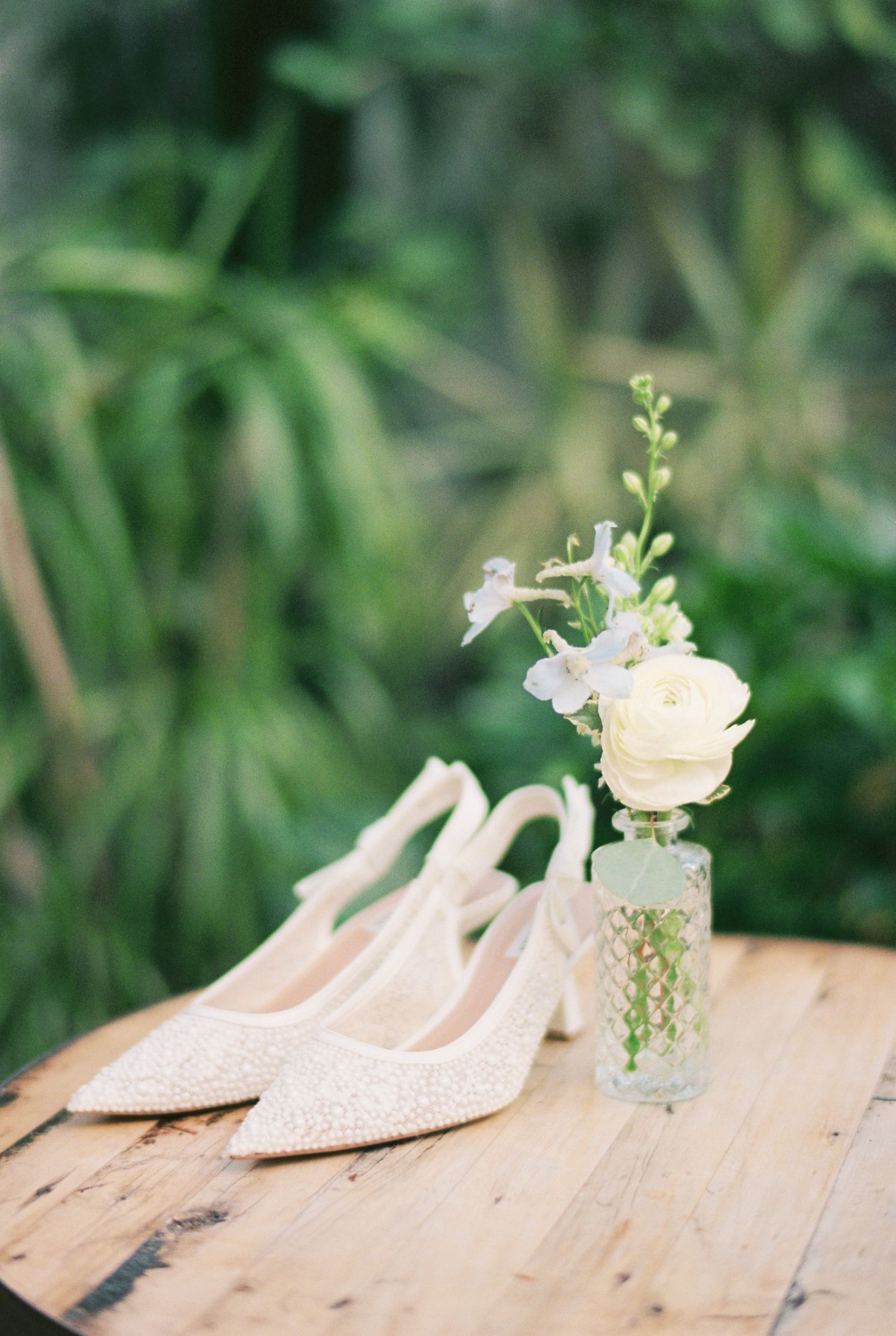 Elegant ivory lace bridal heels beside a delicate floral arrangement in a glass vase on a rustic wooden table.