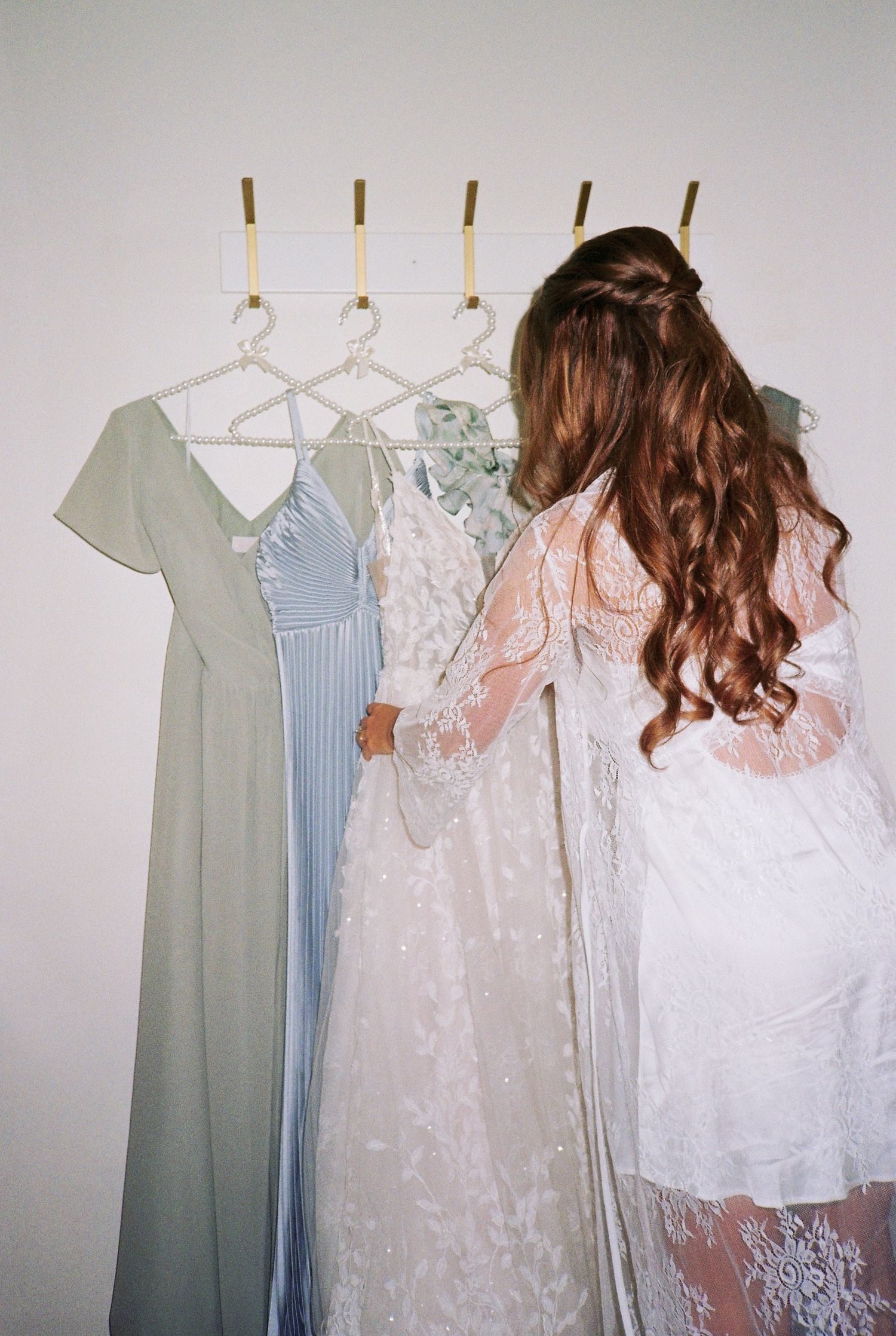 A bride with curly auburn hair views her lace wedding gown and bridesmaid dresses hanging on gold hooks.
