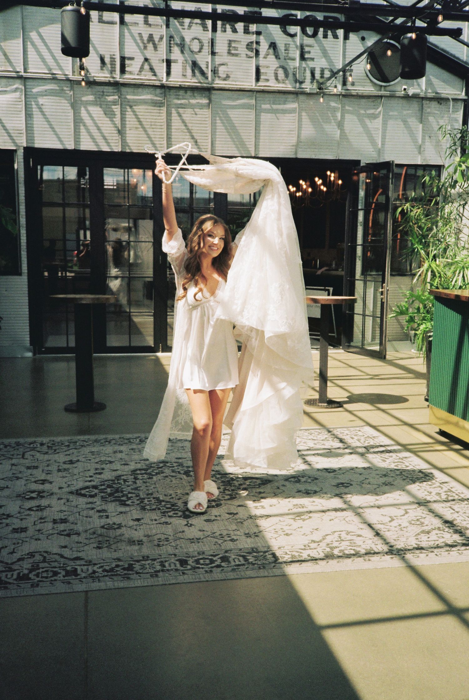Bride in white mini dress twirling wedding gown veil outside modern venue on sunny day.