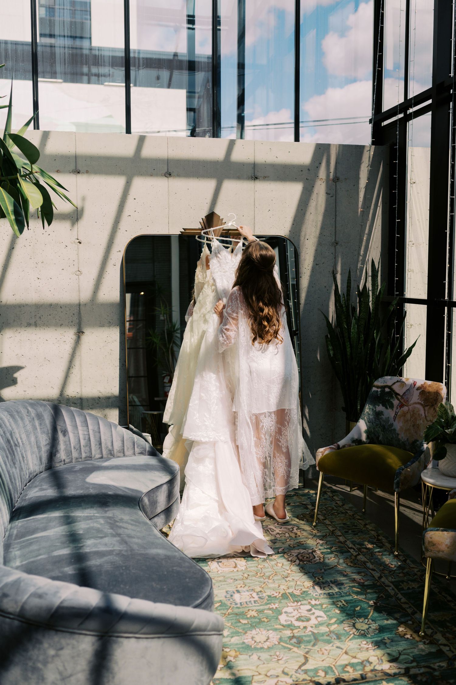Bride holding wedding dress on hangers in a sunlit modern venue with concrete walls and lush greenery.