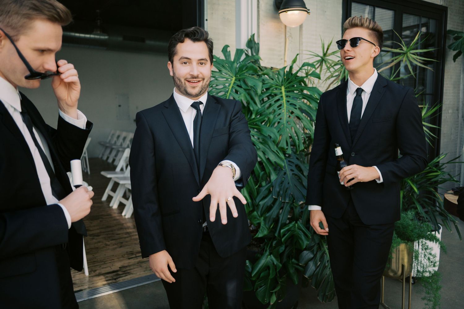 Three men in black suits smiling and gesturing outdoors near tropical plants at a formal event.