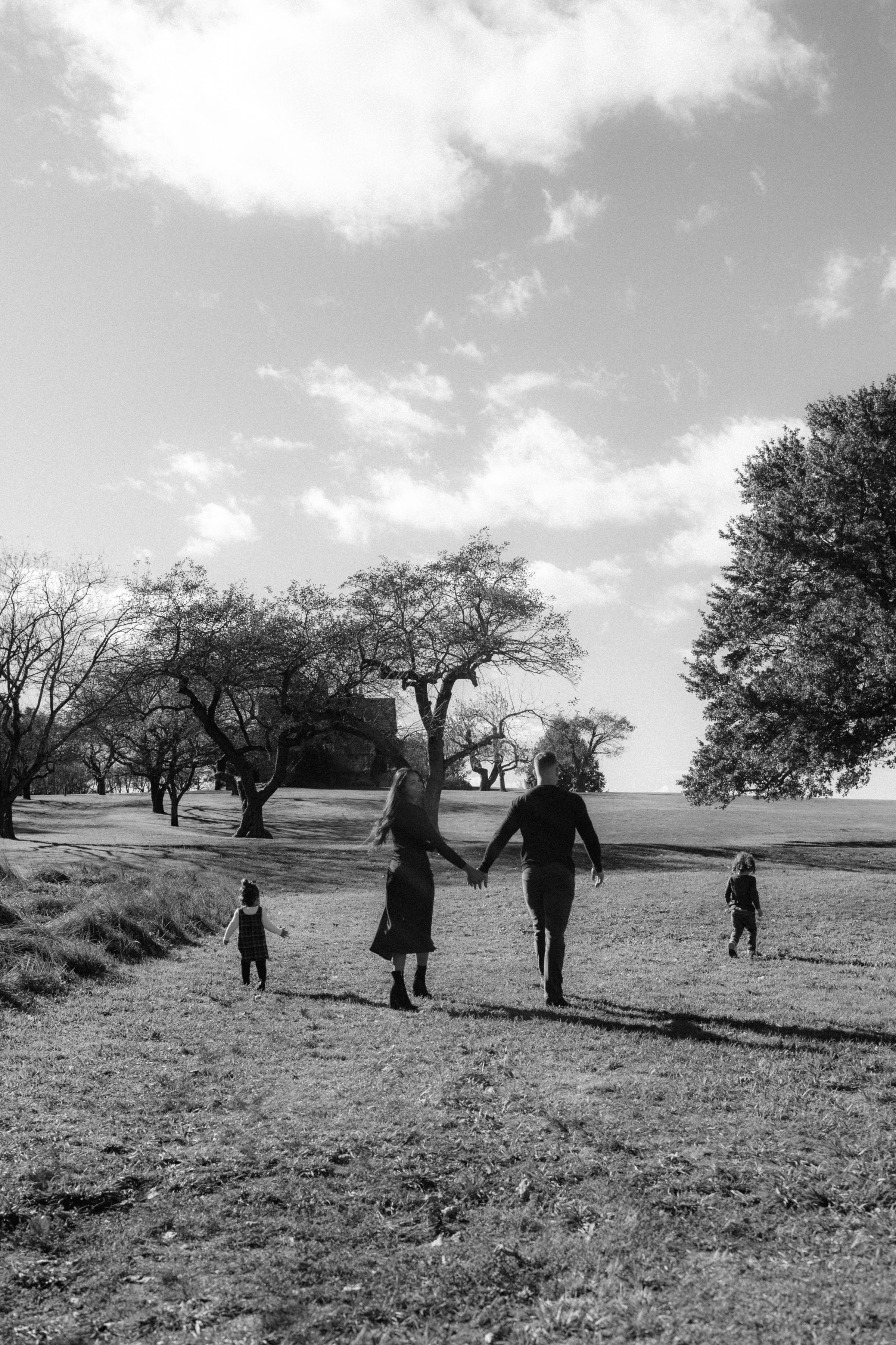 Black and white photo of a family holding hands walking through a scenic park with trees and cloudy sky.