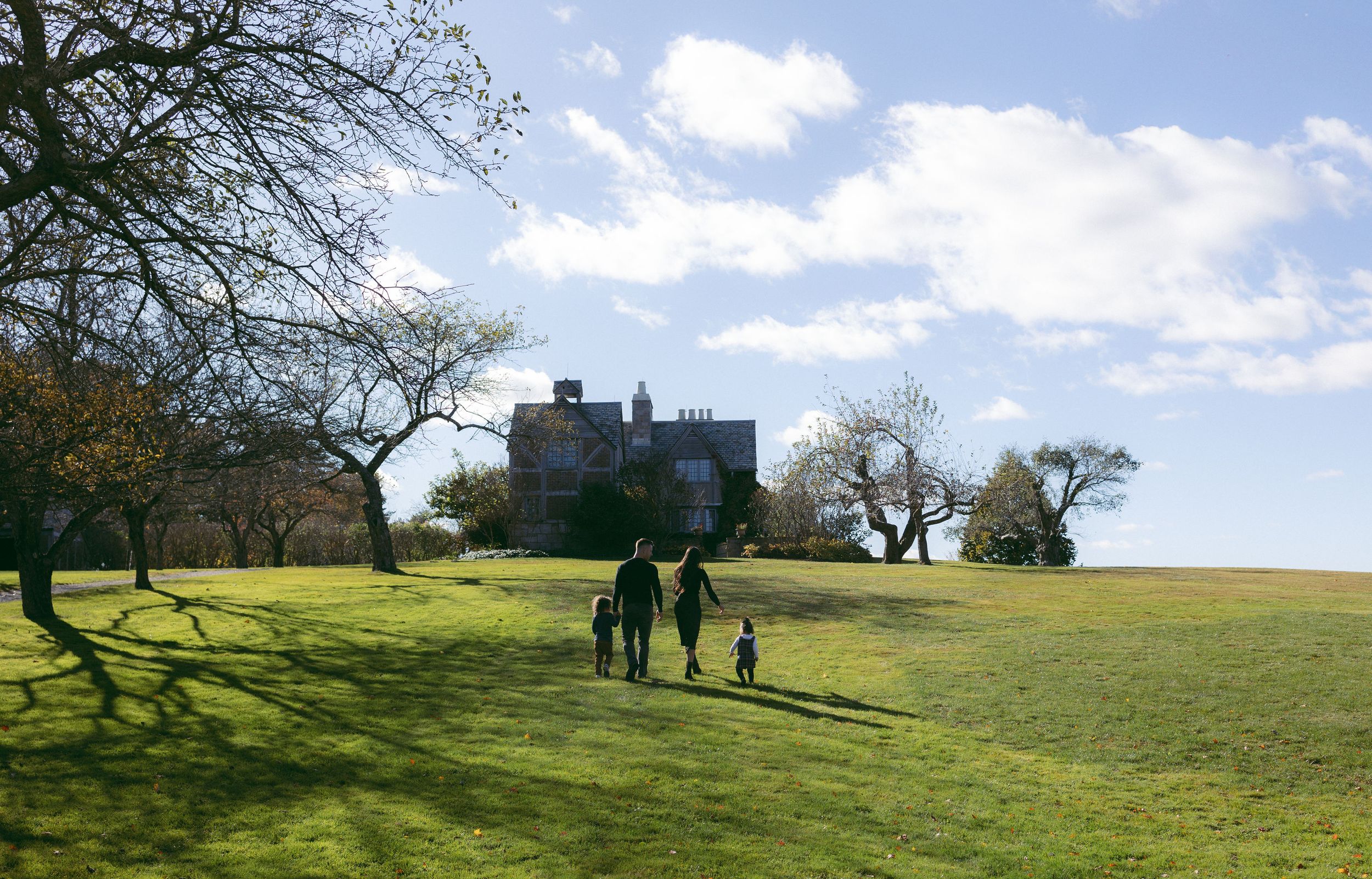 Family walking across green lawns toward a historic manor house on a sunny day with blue skies and bare trees.
