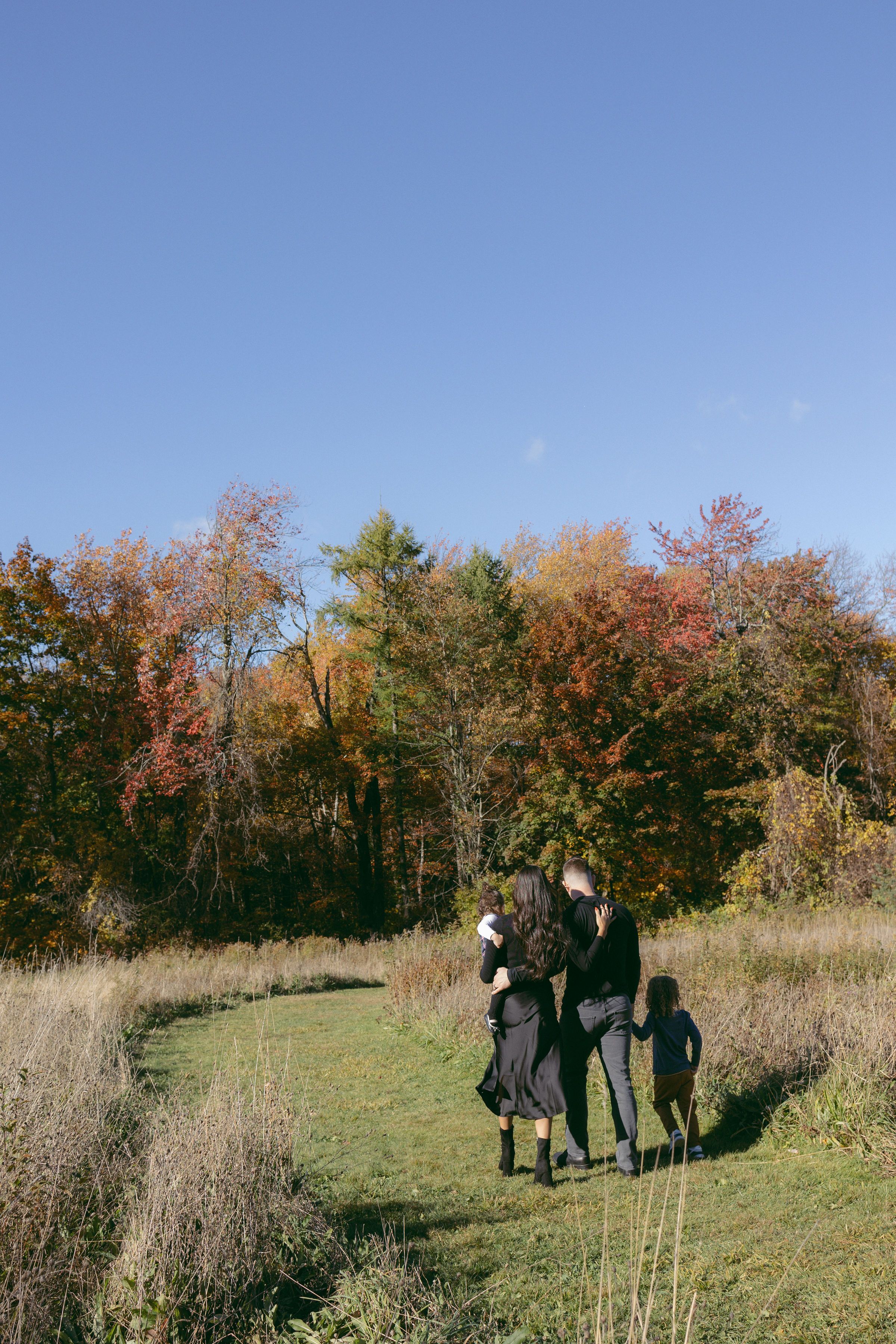 Family walking together on a grassy trail surrounded by colorful autumn trees under a clear blue sky.