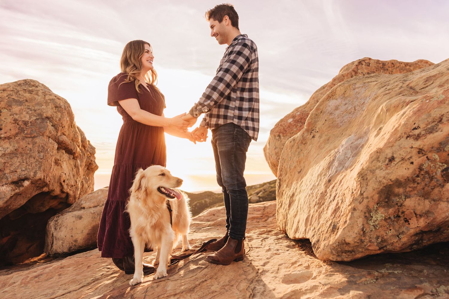 Couple holding hands at golden hour on rocky terrain with a golden retriever dog beside them.