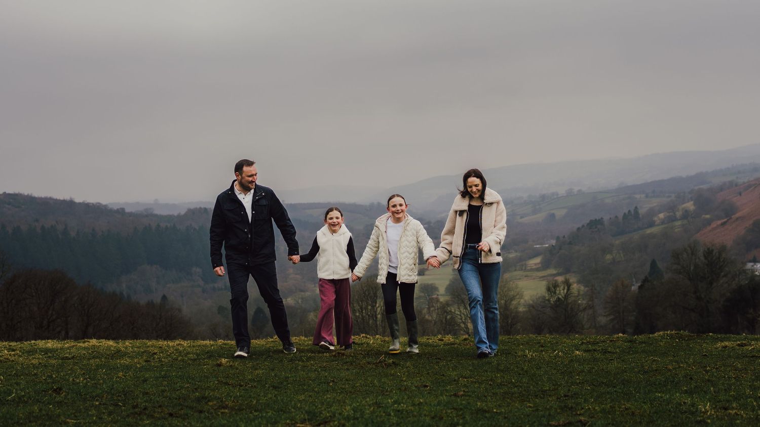 Family of four holding hands on a green hilltop with scenic valley and overcast sky in background.
