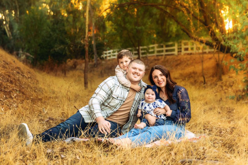 A family sits together on golden grass during sunset with wooden fence and trees in background taken by local photographer in Fresno,ca