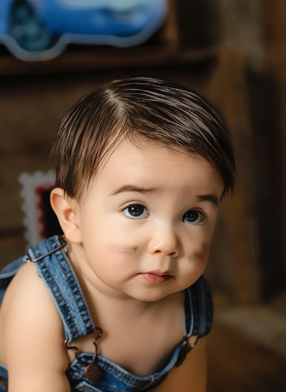 Adorable baby wearing denim overalls looks at camera with bright blue eyes and sweet expression on their face taken by local photographer.