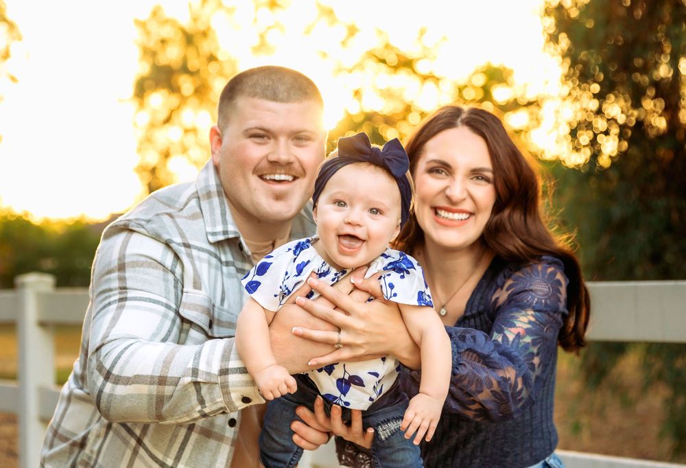 A happy family poses together outdoors at sunset with a white fence in the background taken by local photographer in Fresno,ca