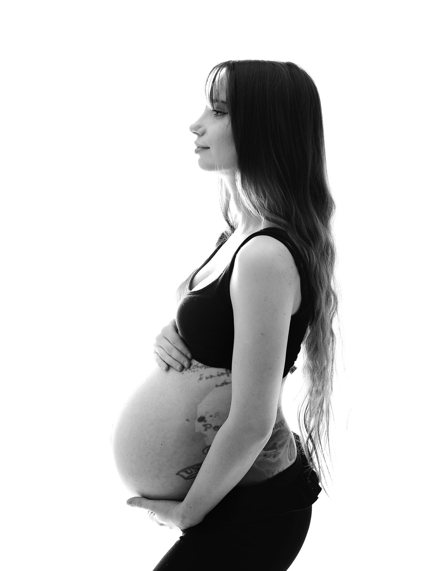 Black and white profile photo of a pregnant woman cradling her baby bump against a white background. During a maternity photoshoot in Bexley