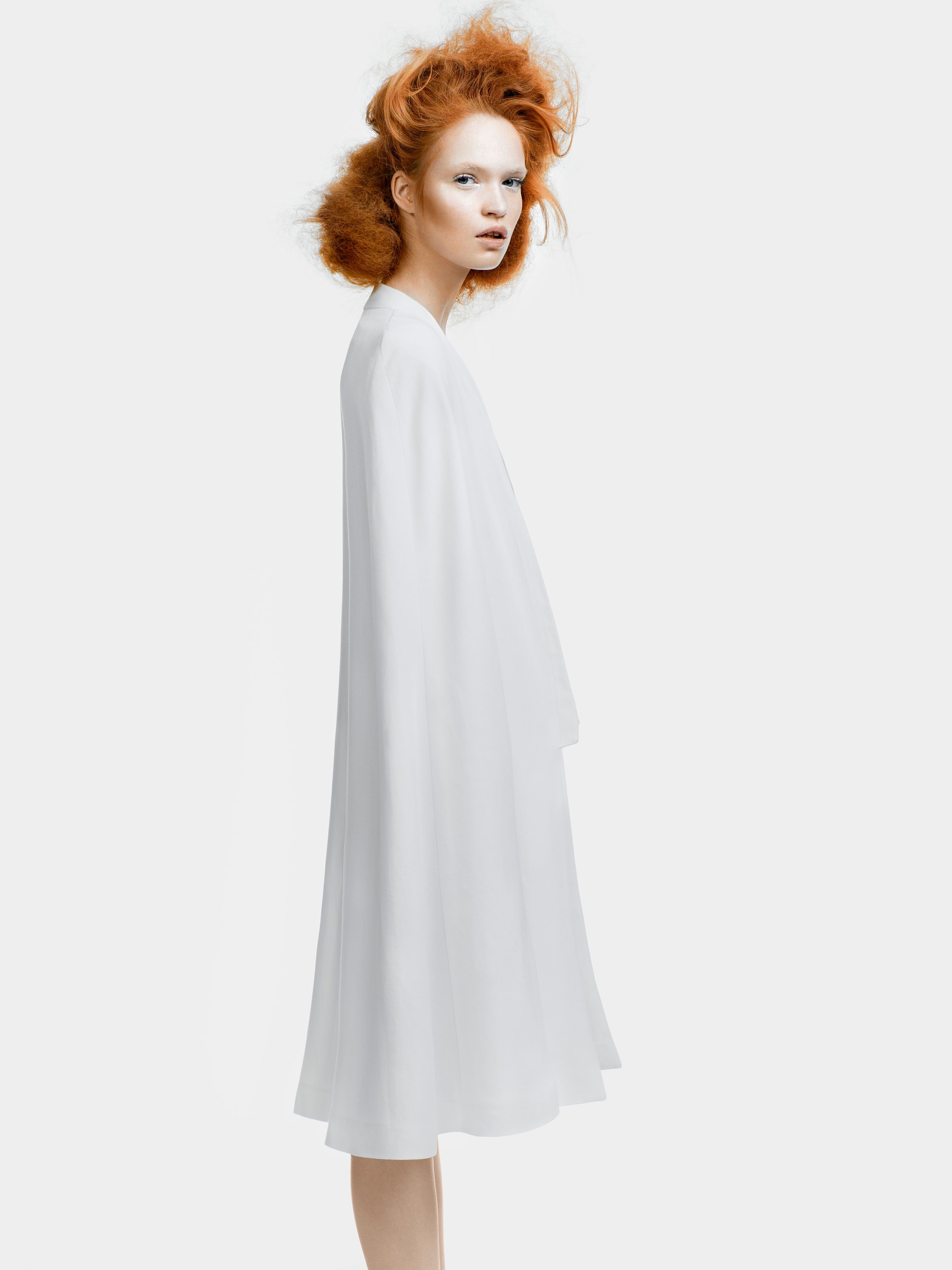 Woman with red updo hair wearing a flowing white midi dress with cape sleeves on white background.