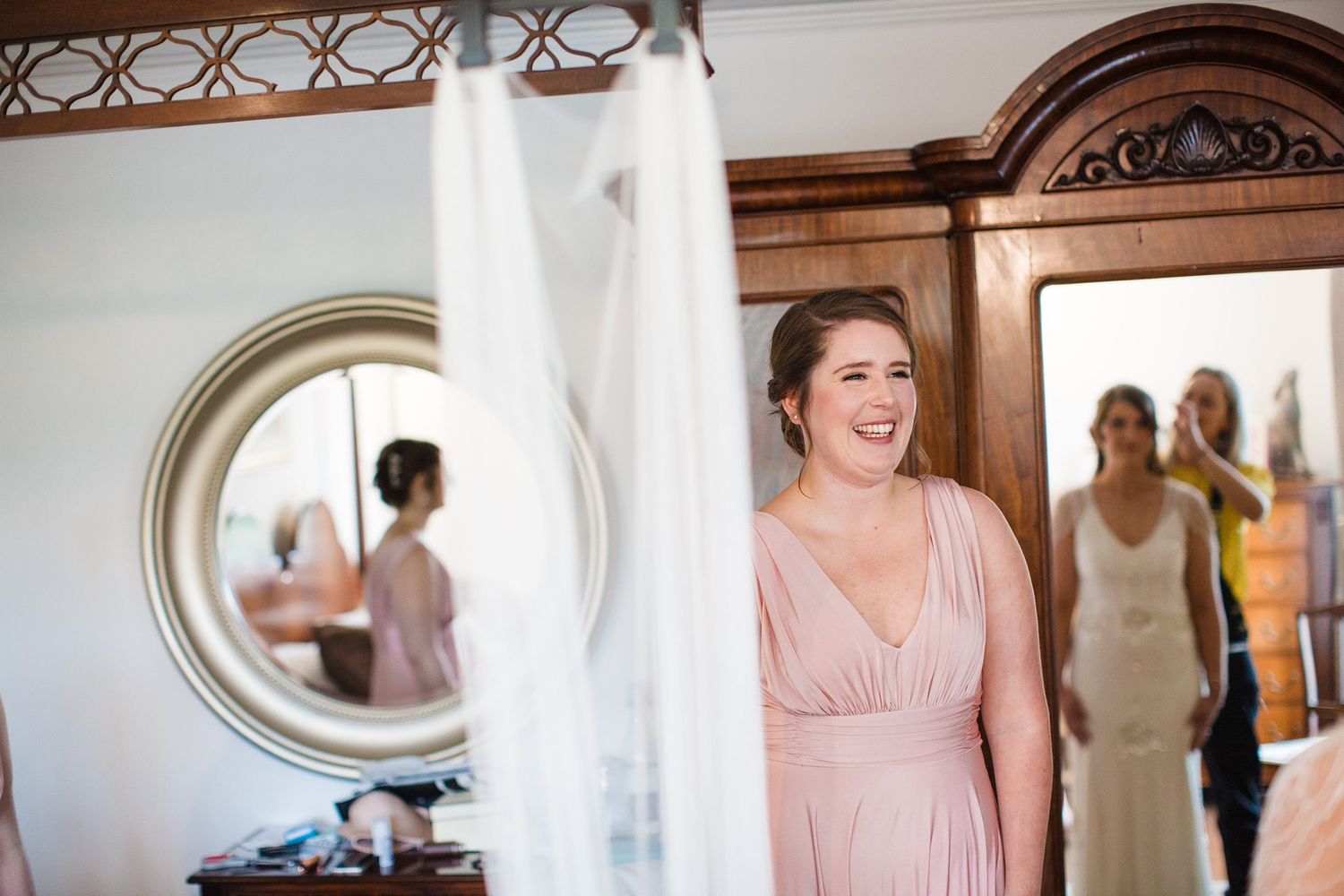 Bridesmaid in pink dress smiling during wedding preparations with bridal gown hanging nearby and mirror reflection in background.