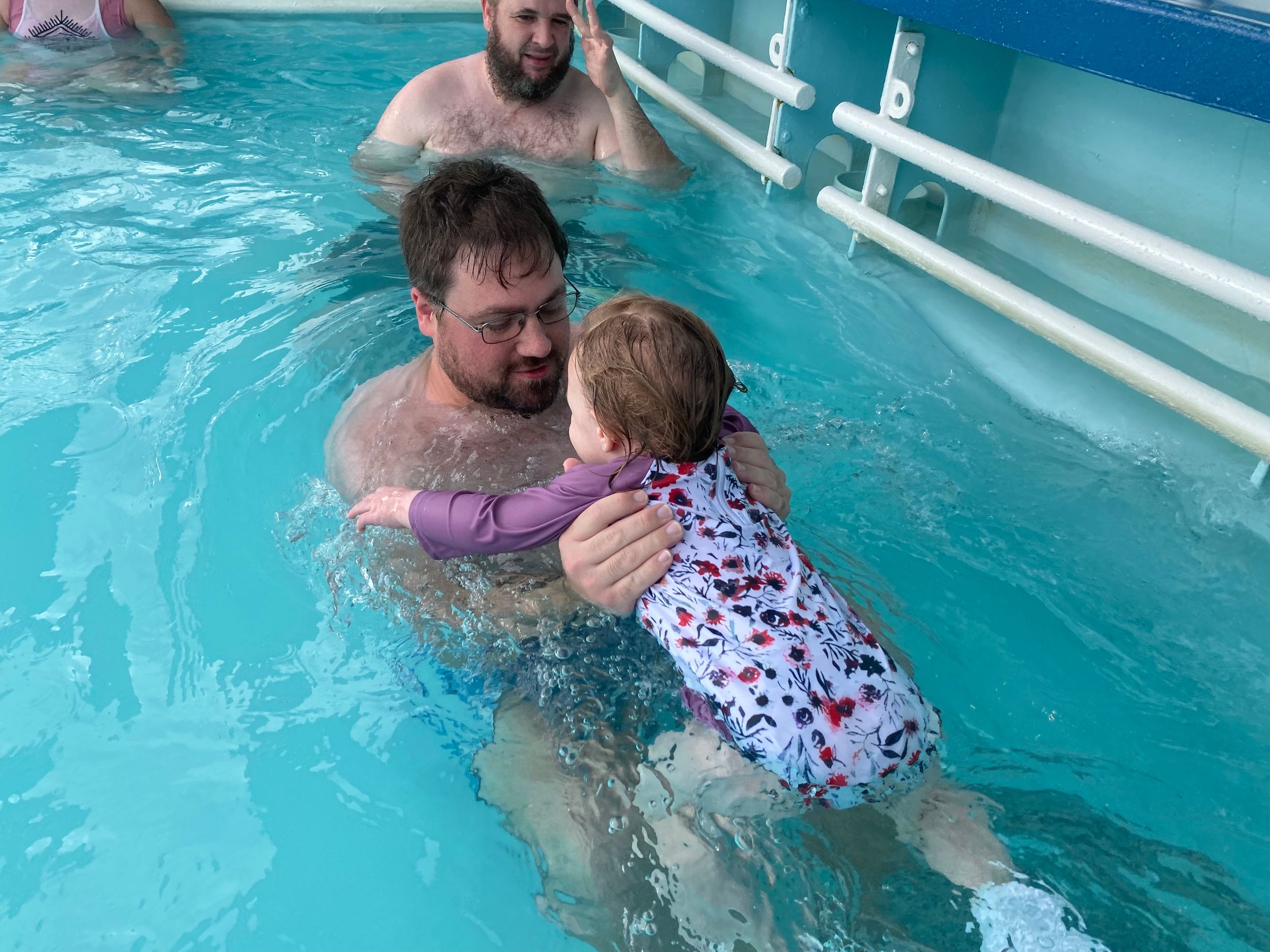 Adult man teaching a toddler to swim in an indoor pool, holding the child in colorful swimwear.