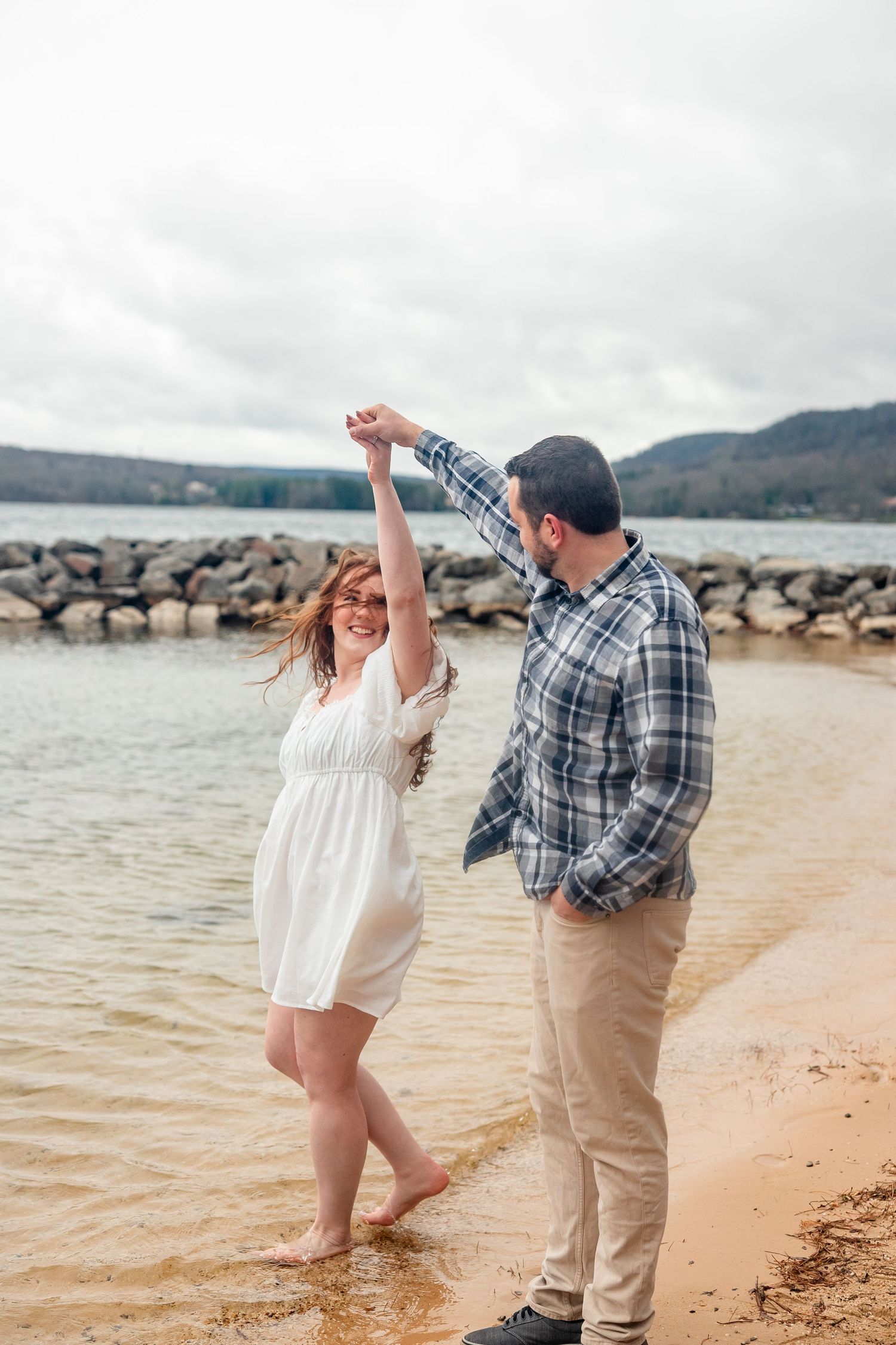 Couple dancing on sandy beach shore, woman in white dress spinning, rocky waterfront and hills in cloudy background. deep creek lake md