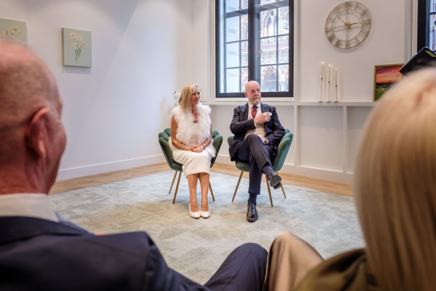 Couple seated in green chairs during an intimate indoor wedding ceremony with guests in foreground.