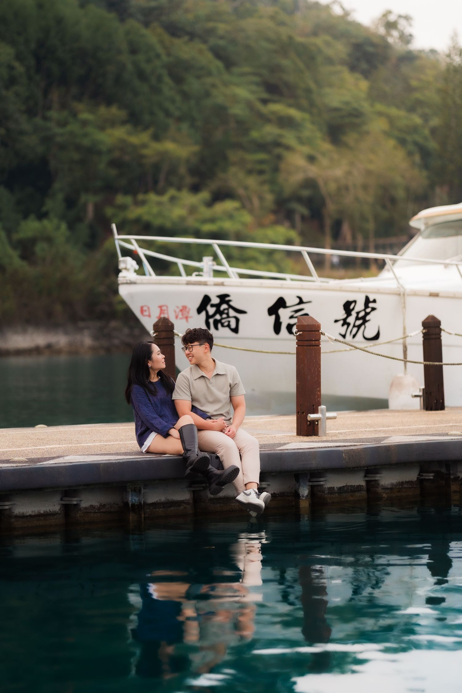 Couple holding hands walking on a pier between boats with misty mountains and calm blue lake in background.