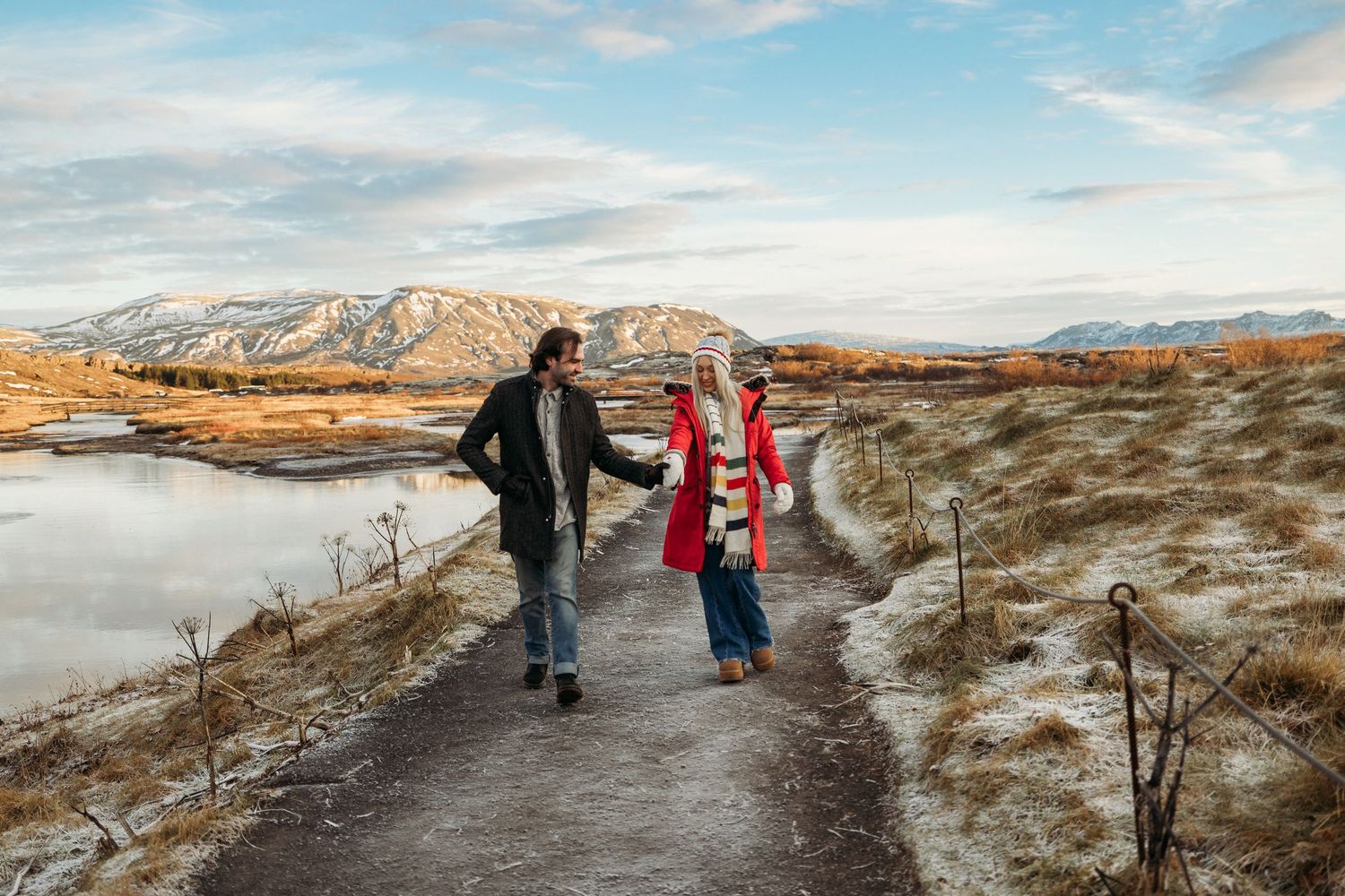 Couple holding hands walking on a winter path by a frozen lake with snow-dusted mountains in the background.