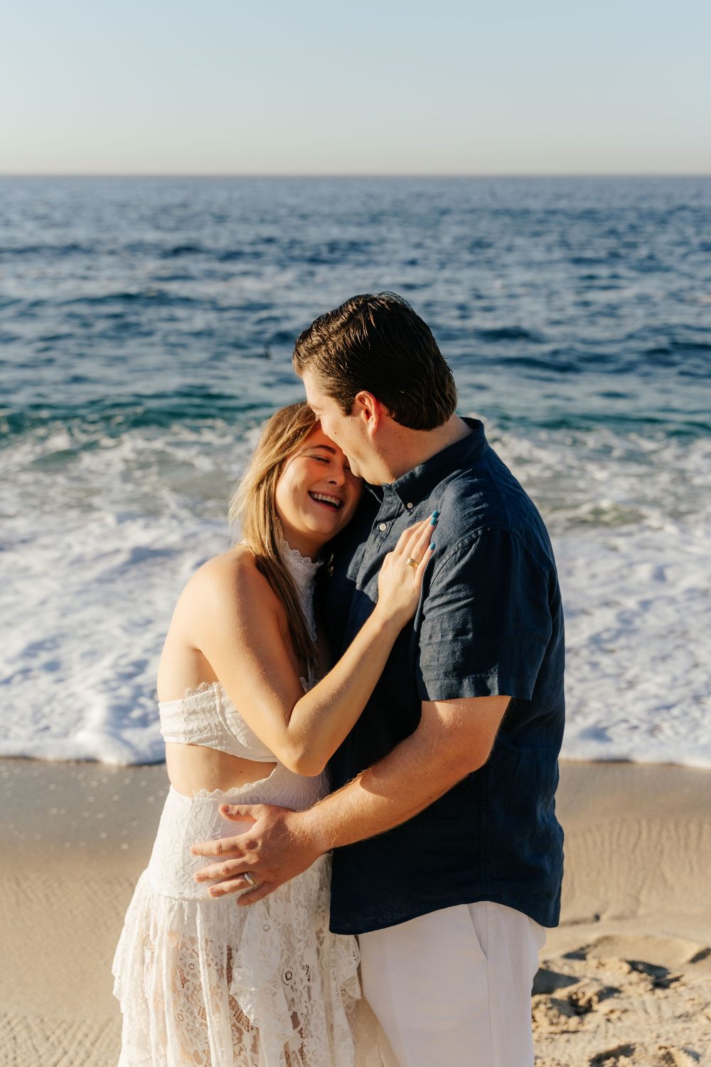 A couple embraces and laughs together on a sandy beach with ocean waves behind them.