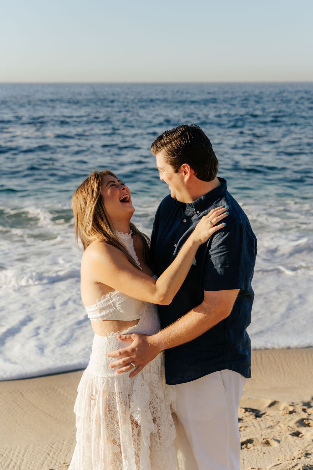 A laughing couple embraces on a sandy beach with ocean waves behind them, woman in white lace dress, man in navy shirt.
