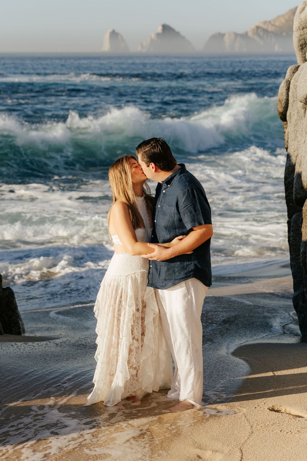 Couple kissing on sandy beach with ocean waves and rocky cliffs in Cabo San Lucas at golden hour.