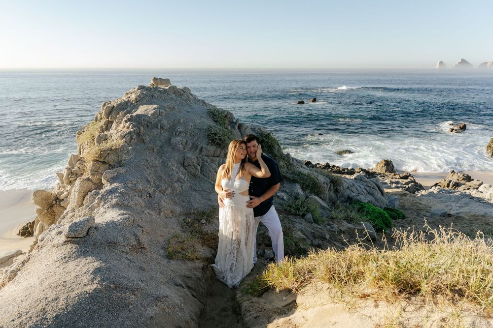 Couple embracing on rocky coastal cliffs with ocean waves and clear blue sky in the background.