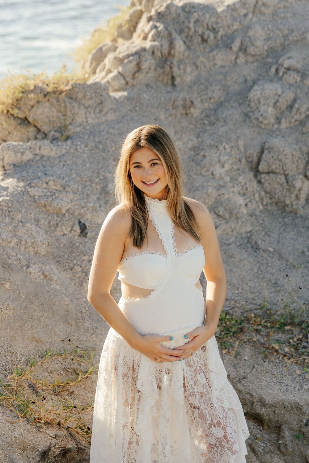 Smiling pregnant woman in white lace halter gown cradling baby bump outdoors near rocky coastal scenery in golden light.
