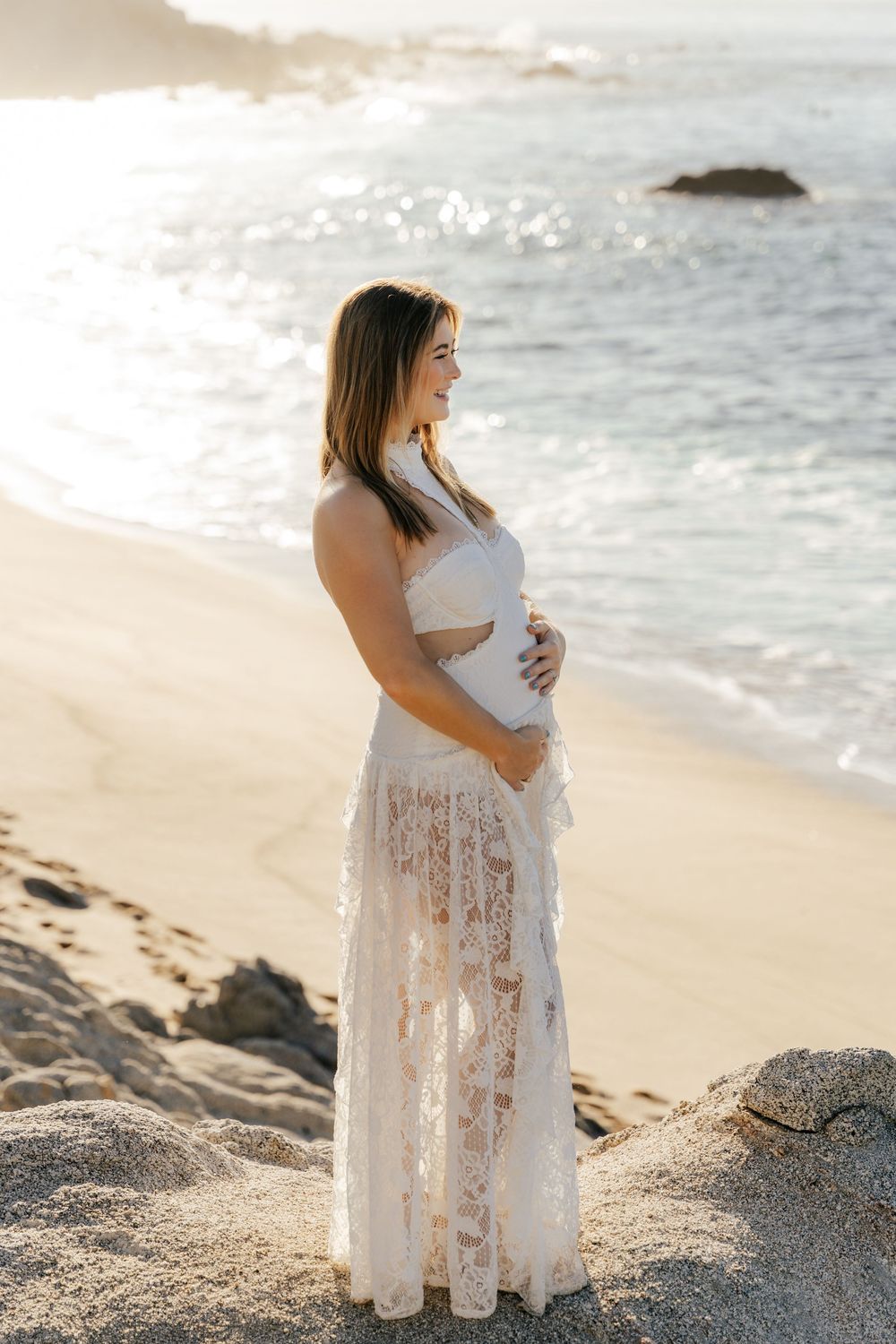 Pregnant woman in white lace dress cradling baby bump on sunny beach at golden hour.