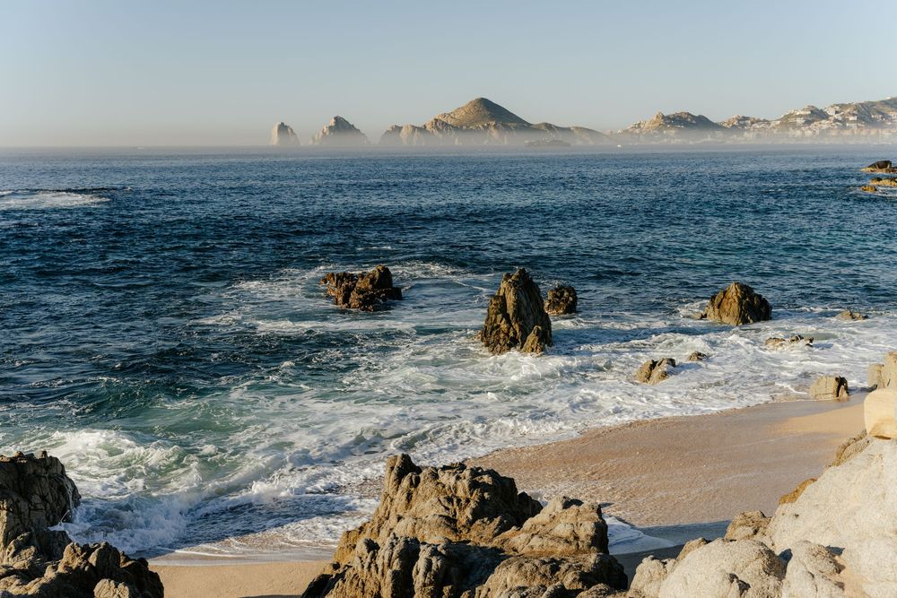 Rocky shoreline with crashing waves and misty mountain peaks in the background at a scenic coastal beach.