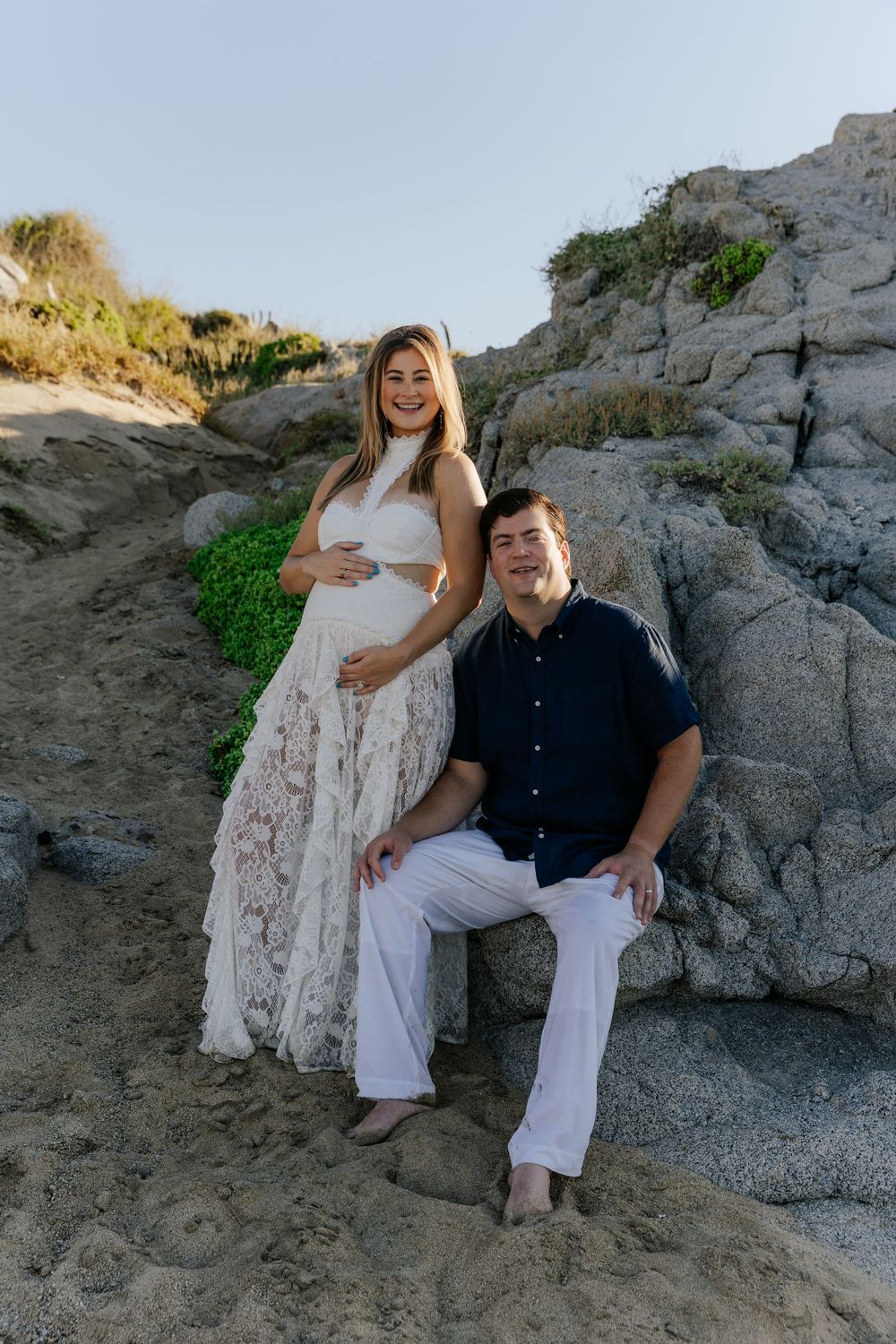 Pregnant woman in white lace gown poses with man in navy shirt on rocky beach during maternity photo session.