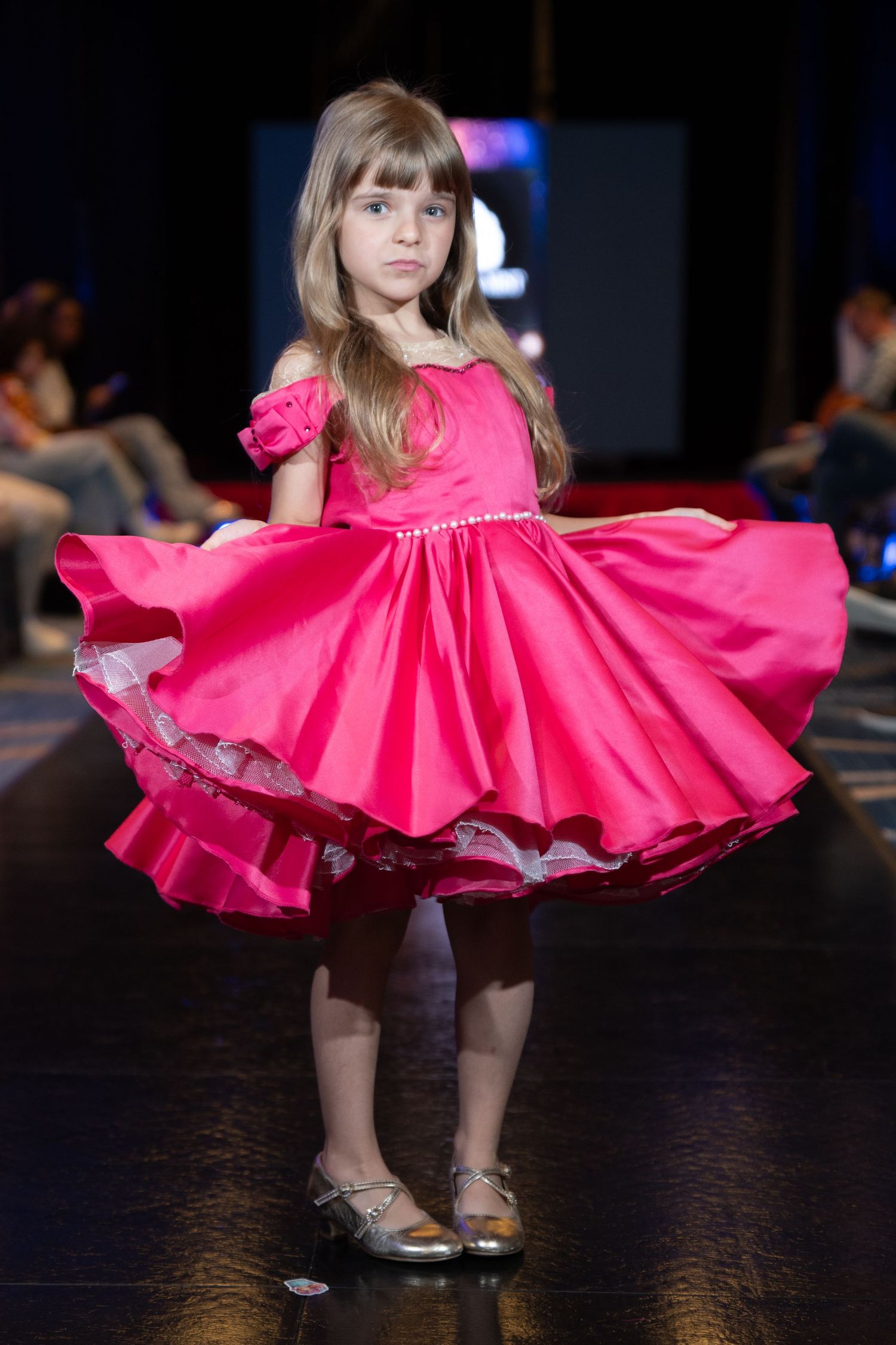Young girl modeling an elegant red and pink floral ruffled gown on a fashion runway.