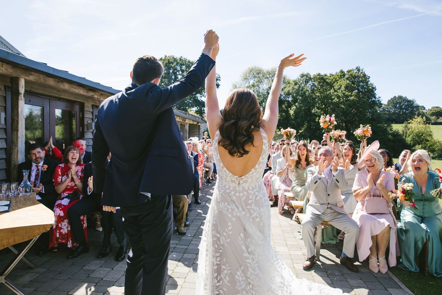 Bride and groom raising arms in celebration before cheering wedding guests outdoors on a sunny day.