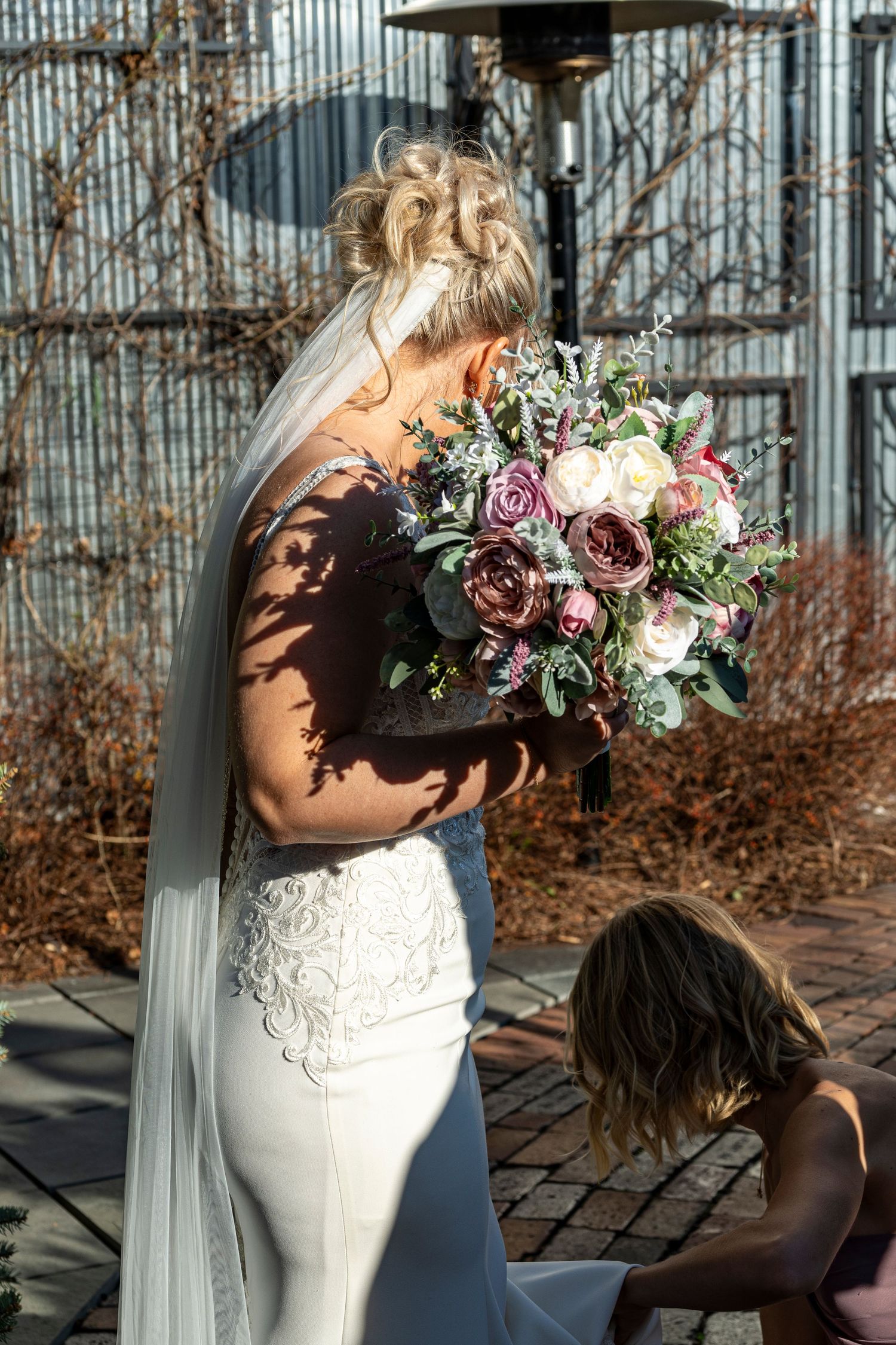 Bride in white lace gown holding purple and pink floral bouquet while flower girl adjusts her dress outdoors.