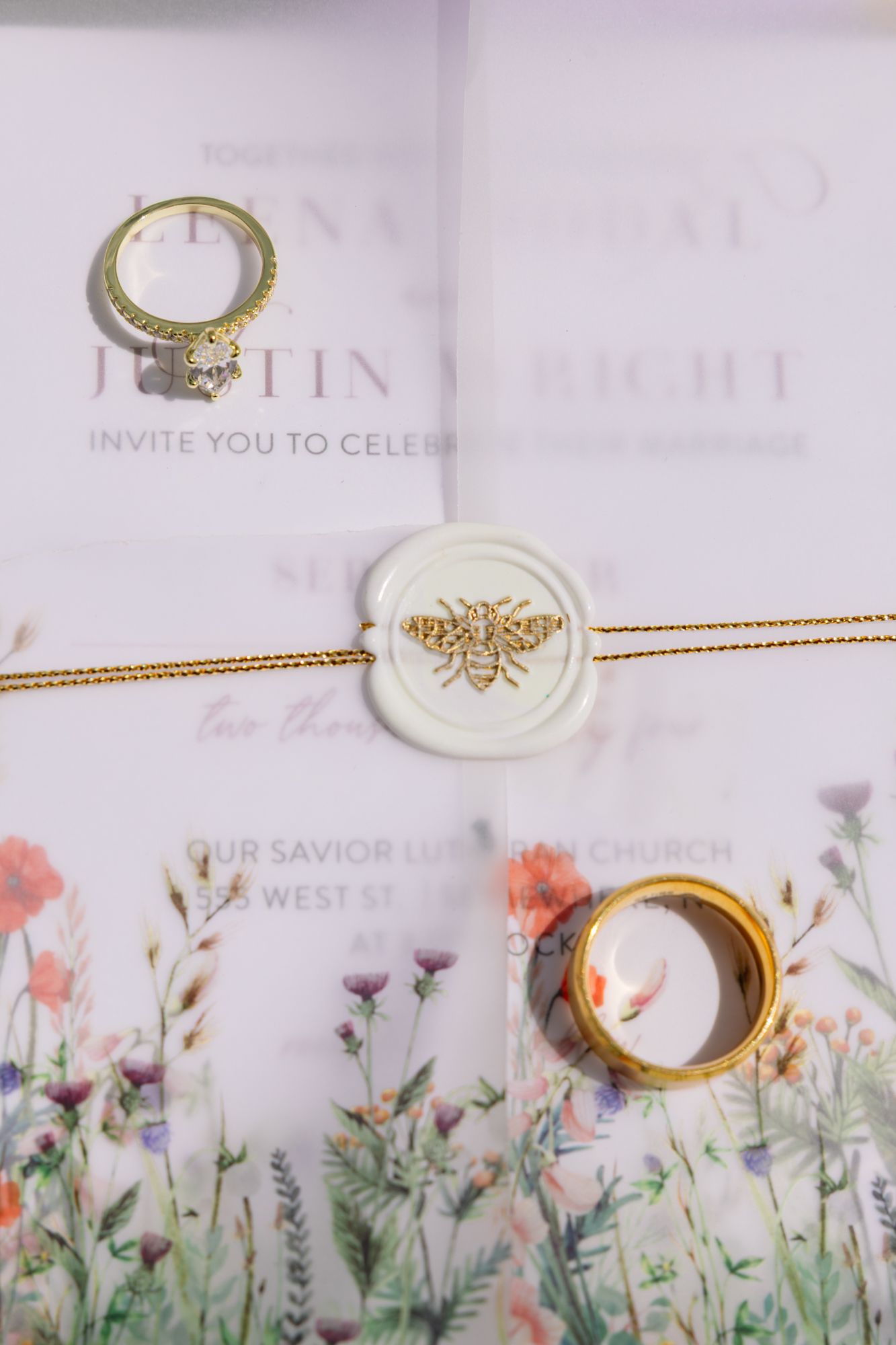Wedding invitation with floral design features gold rings and a decorative wax seal with bee emblem on vellum paper.