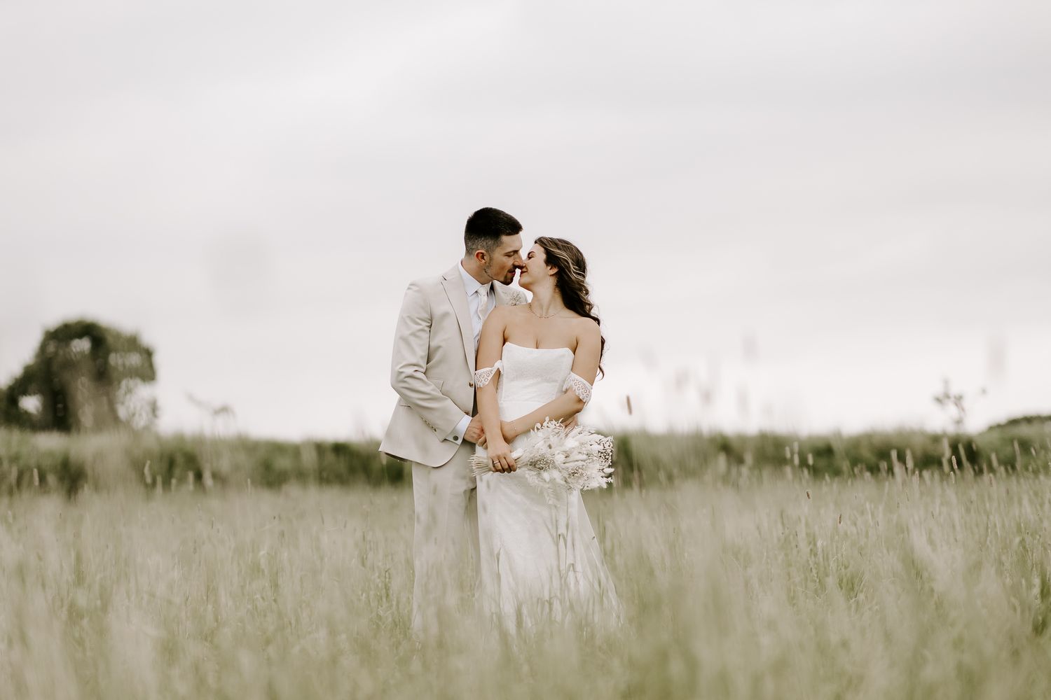 A romantic couple embraces in a serene meadow with tall grass under an overcast sky.