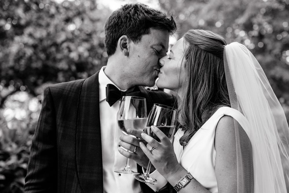 Black-and-white portrait of a British bride and groom kissing while holding champagne glasses at Château de Sainte-Croix in the Dordogne, Fr