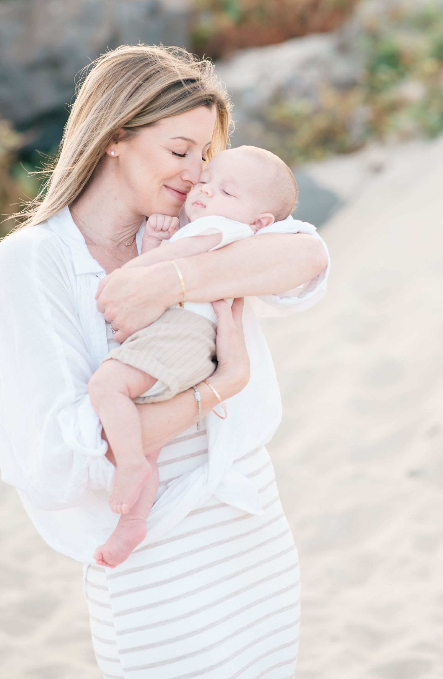A young mother with blond hair and a dress pulls her baby boy in close and tickles his face with her nose.